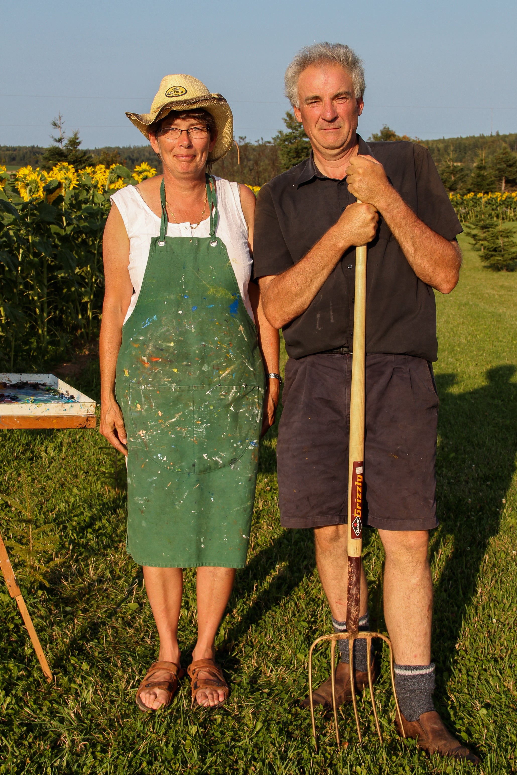 Two people, one in a green apron and the other holding a garden fork, standing in a field with sunflowers.