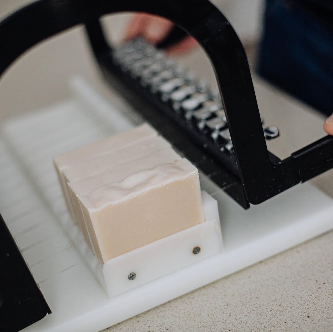 Close-up of a soap bar being sliced with a soap cutter on a white surface.