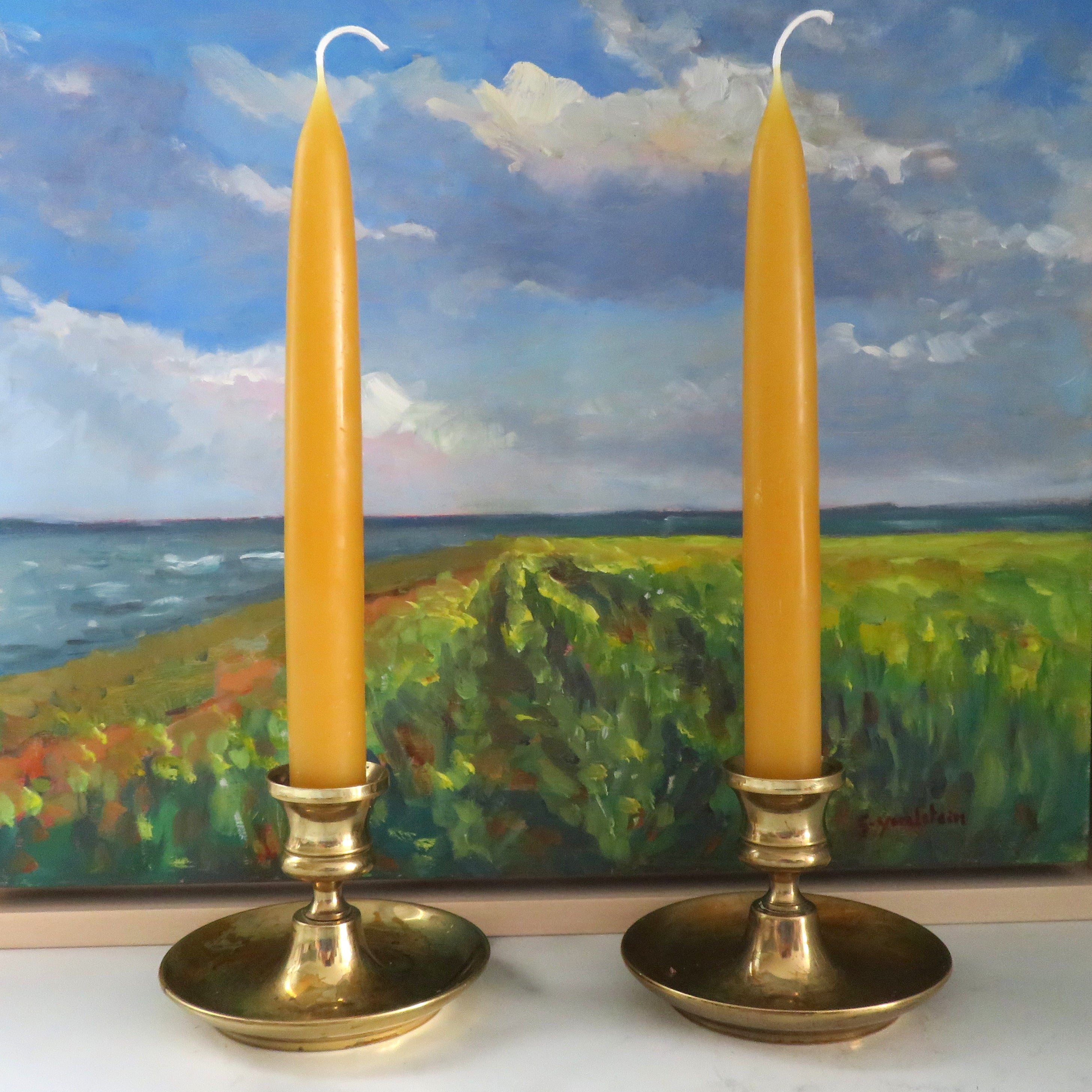 Two yellow candles in brass candle holders with a scenic painting in the background.