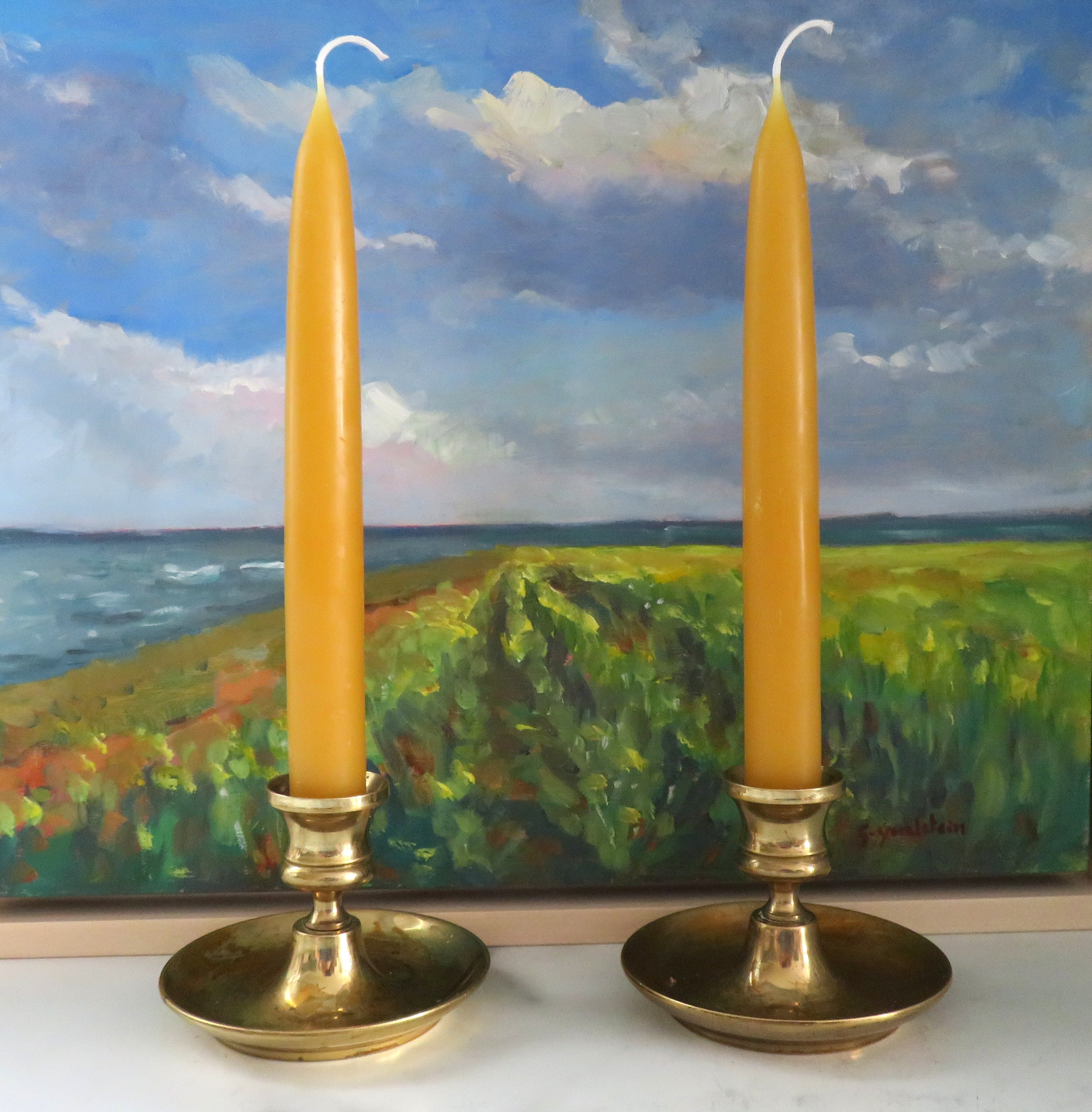 Two yellow candles in brass candle holders with a scenic painting in the background.