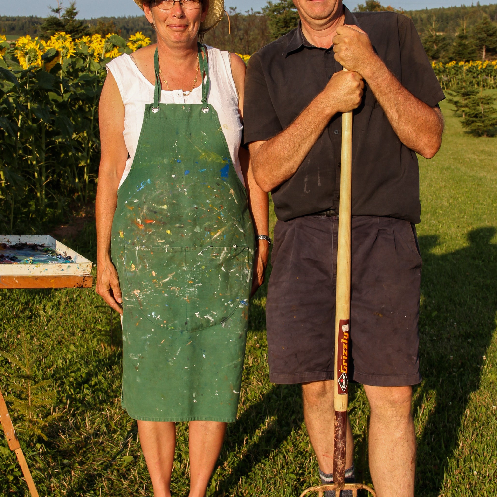 Two people, one in a green apron and the other holding a garden fork, standing in a field with sunflowers.