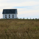 Small white church with a black roof in a grassy field under a clear sky.
