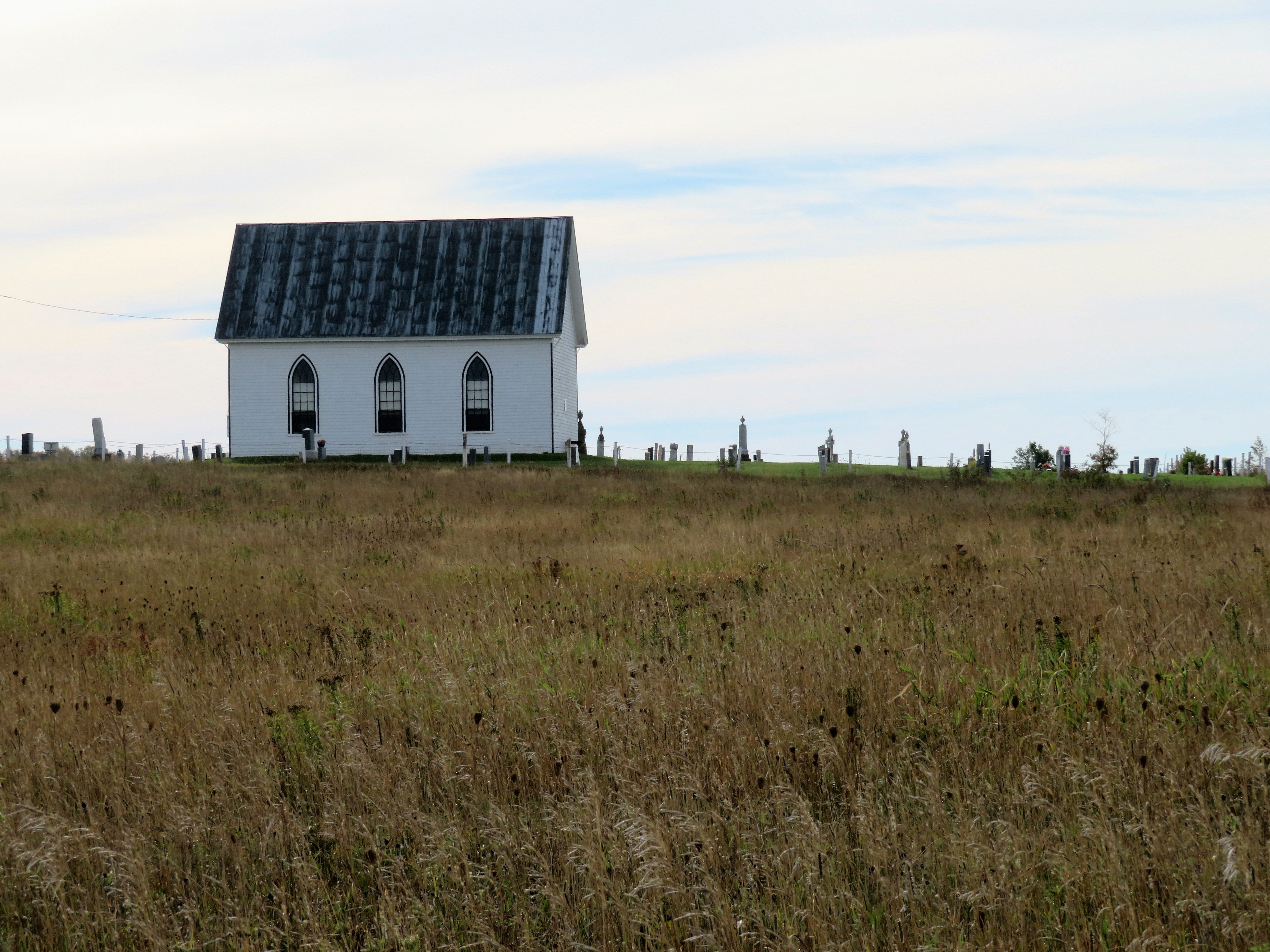 Small white church with a black roof in a grassy field under a clear sky.