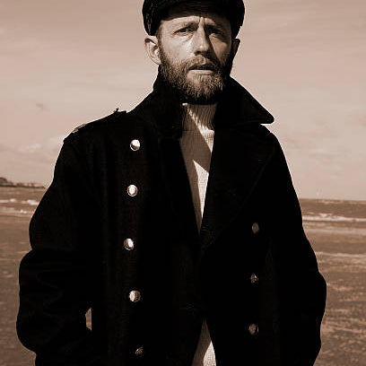 Man wearing a dark coat and hat in a sepia-toned landscape