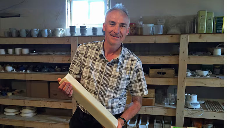 Man holding a ceramic piece in a pottery studio with shelves of pottery in the background